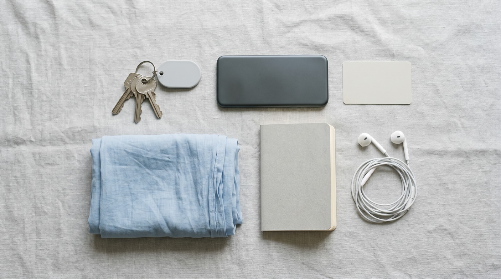 Everyday carry laid out on grey linen: house keys with a small grey key-ring fob, a phone, a blue dupatta, a metro card, a notebook and earphones