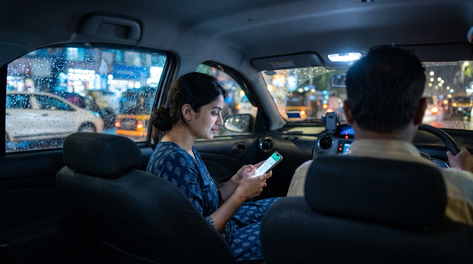 A woman in the back seat of a taxi at night following the route on her phone as the driver drives